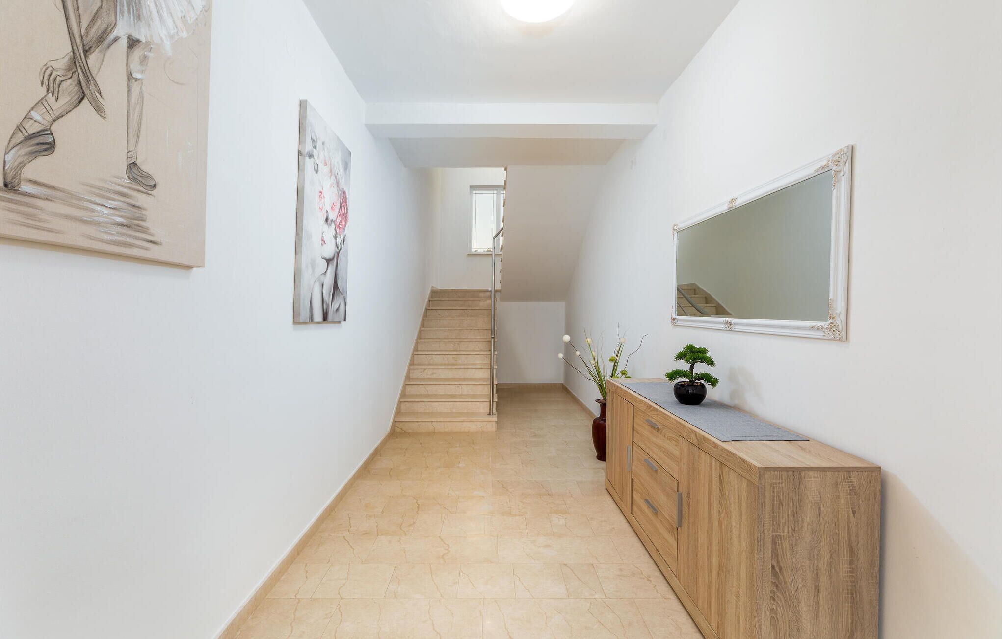 A hallway with a staircase, a wooden cabinet, a mirror, and a painting on the wall.