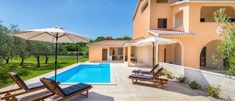 A poolside area with lounge chairs and umbrellas, a swimming pool, and a building with a tiled roof.
