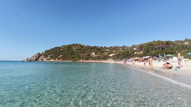 Una spiaggia con acque cristalline, persone che prendono il sole e una collina con edifici sullo sfondo.