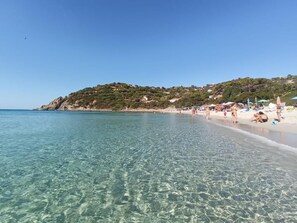 A beach with clear water, people sunbathing, and a hill with buildings in the background. - A cozy home just a stone's throw from the sea (Torre Delle Stelle (Maracalagonis))