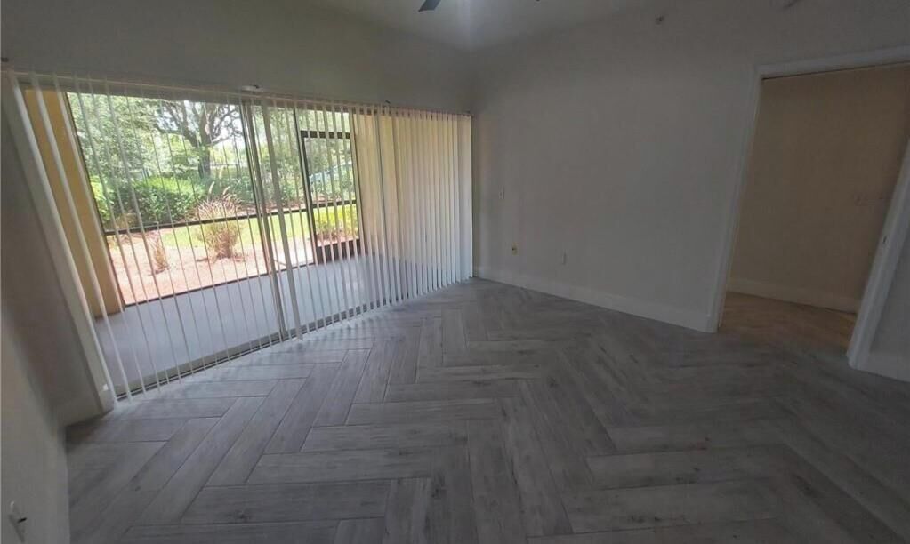 A room with a herringbone-patterned wooden floor, a ceiling fan, and a large window with curtains.