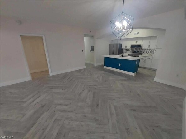 A spacious living area with a herringbone-patterned floor, a central island with a blue countertop, and a modern kitchen with white cabinets and a hexagonal pendant light. - GORGEOUS TOTALLY RENOVATED 3BED,2BATH CONDO IN GREAT COMMUNITY!!! (Naples)