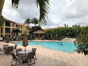 A hotel pool area with lounge chairs and umbrellas, surrounded by palm trees and a building with a red roof. - GORGEOUS TOTALLY RENOVATED 3BED,2BATH CONDO IN GREAT COMMUNITY!!! (Naples)