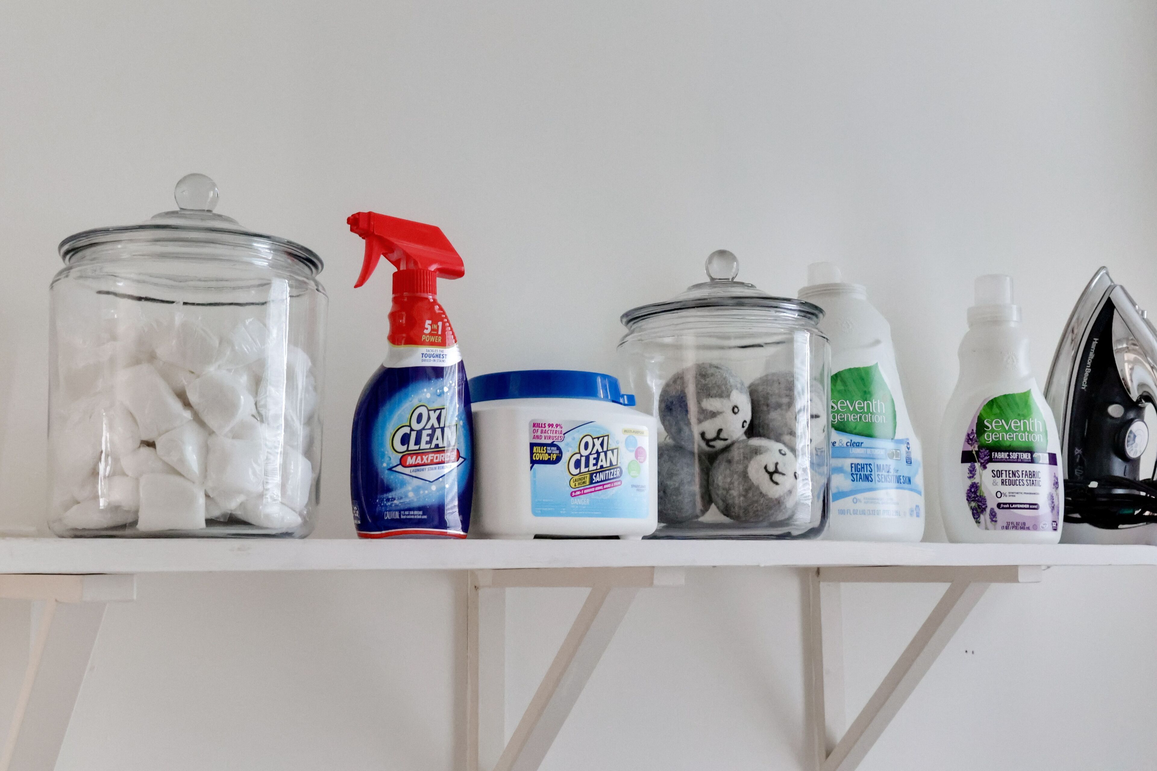 A shelf with cleaning supplies including a spray bottle, a container of fabric softener, and a jar of cotton balls.