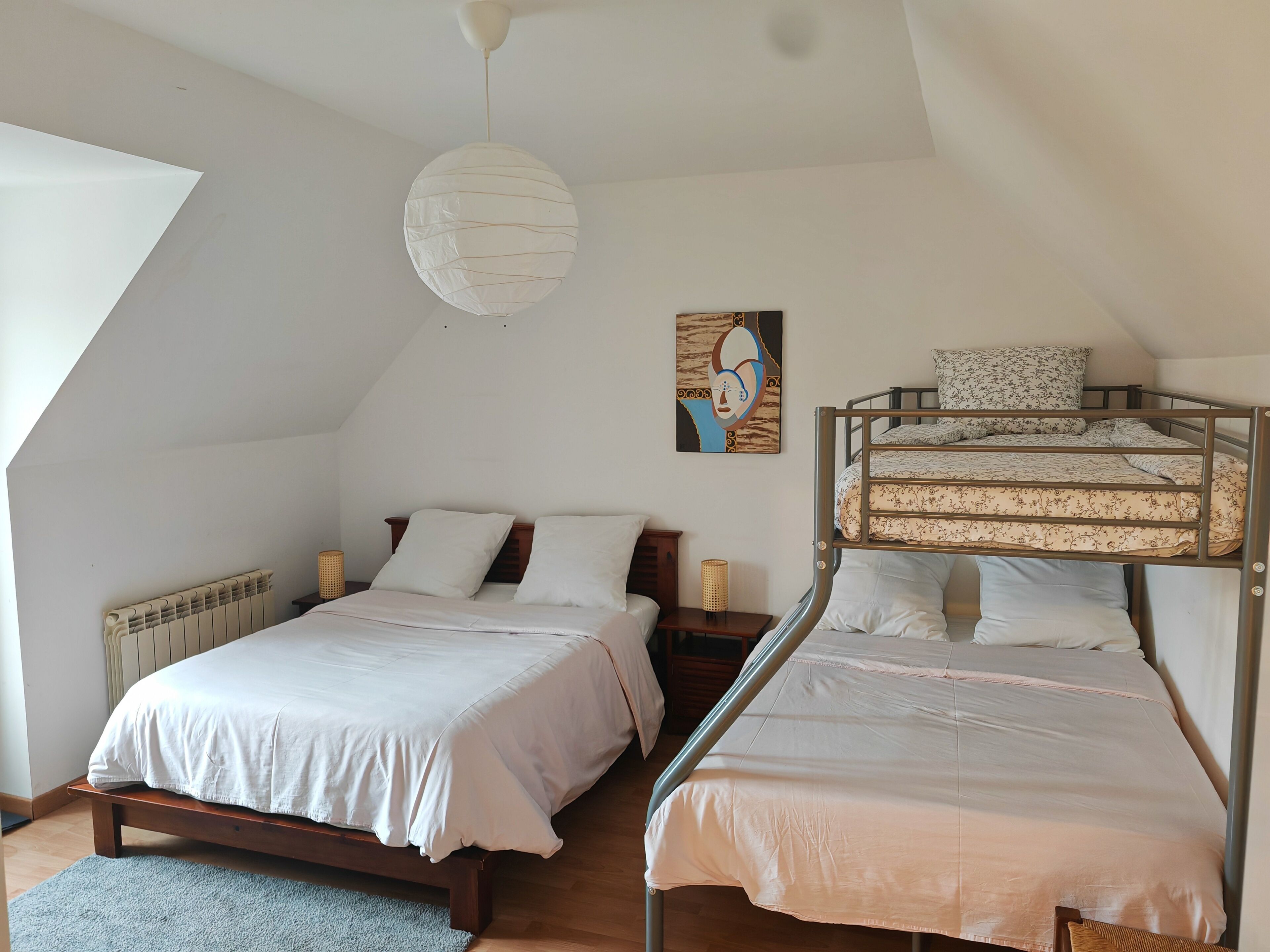 A bedroom with a sloped ceiling, a bunk bed, a single bed, a radiator, a hanging light fixture, and a painting on the wall.