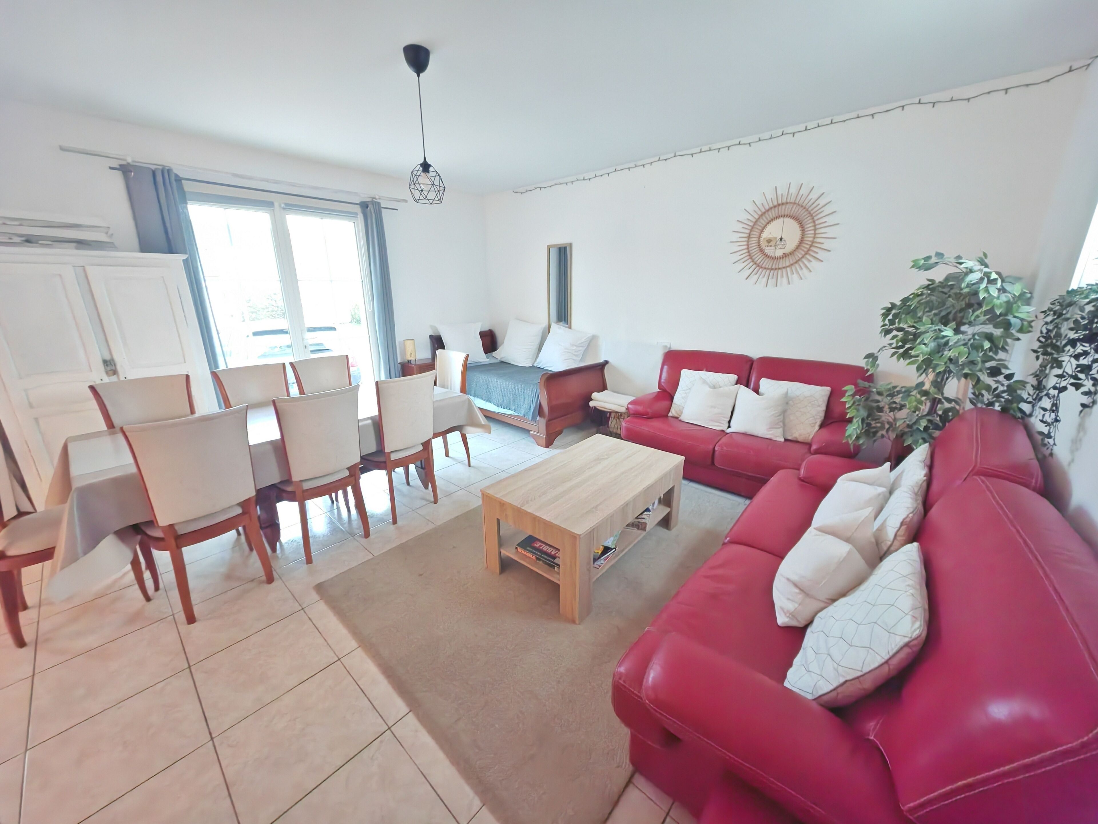 A living room with a red sofa, a wooden coffee table, a dining area with chairs, a bed, and a window with curtains.