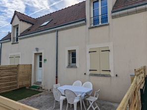 A residential building with a tiled roof, a small balcony, a window with shutters, and a table with chairs outside.