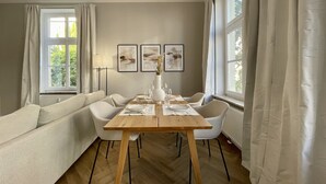 A dining area with a wooden table set for four, surrounded by white chairs, a sofa, and a floor lamp. There are two framed artworks on the wall and a window with curtains on the left. - Apartment Villa Anna - Apartment with parking space (Emden)