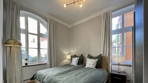 A bedroom with a large bed, two bedside tables, a floor lamp, and a view of a building through a window. - Apartment Villa Anna - Apartment with parking space (Emden)