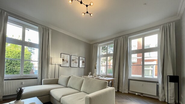 A modern living room with a sectional sofa, large windows, and a herringbone floor. - Apartment Villa Anna - Apartment with parking space (Emden)