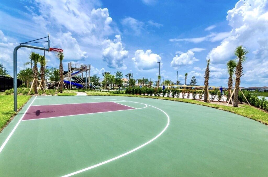 Un terrain de basket en plein air avec une surface de jeu verte et violette, entouré de palmiers et surplombé d’un ciel dégagé.