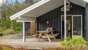 Una terraza de madera con una mesa y sillas de picnic, un espacio de estar cubierto y un edificio de madera oscura con puerta de cristal.