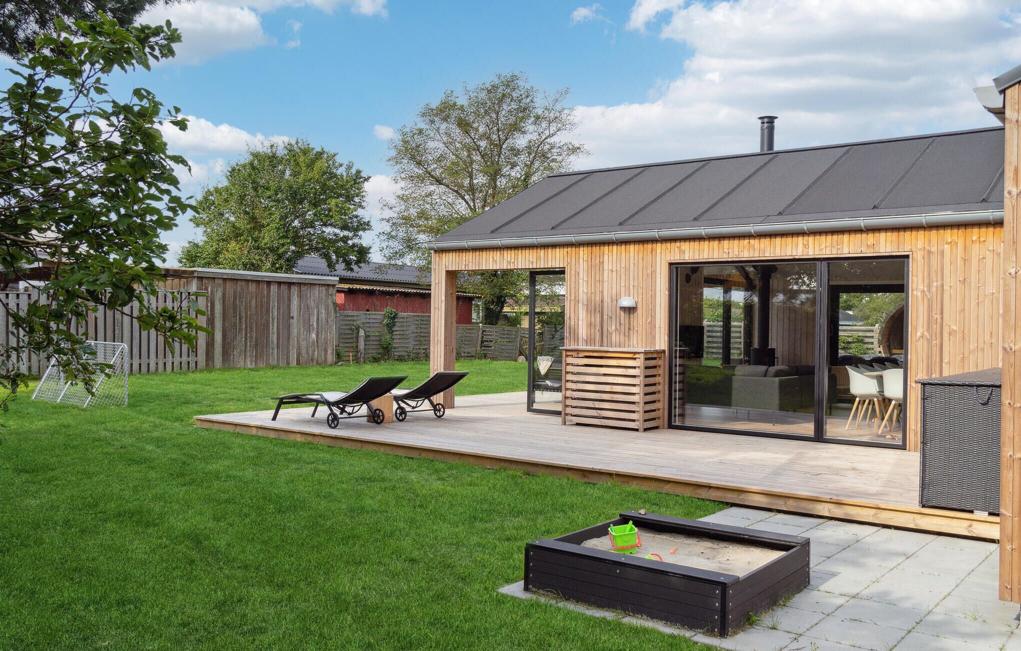 Ein modernes Haus mit Holzfassade, eine Terrasse mit zwei Liegestühlen, ein kleiner Garten und eine große Glasschiebetür zu einem Innenbereich.