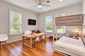 A room with a bunk bed, a desk with chairs, a television mounted on the wall, and large windows with blinds.