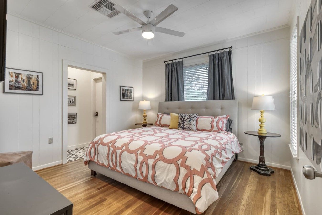 Un dormitorio con cama, dos mesitas de noche con lámparas, un ventilador de techo y una puerta que da a otra habitación.