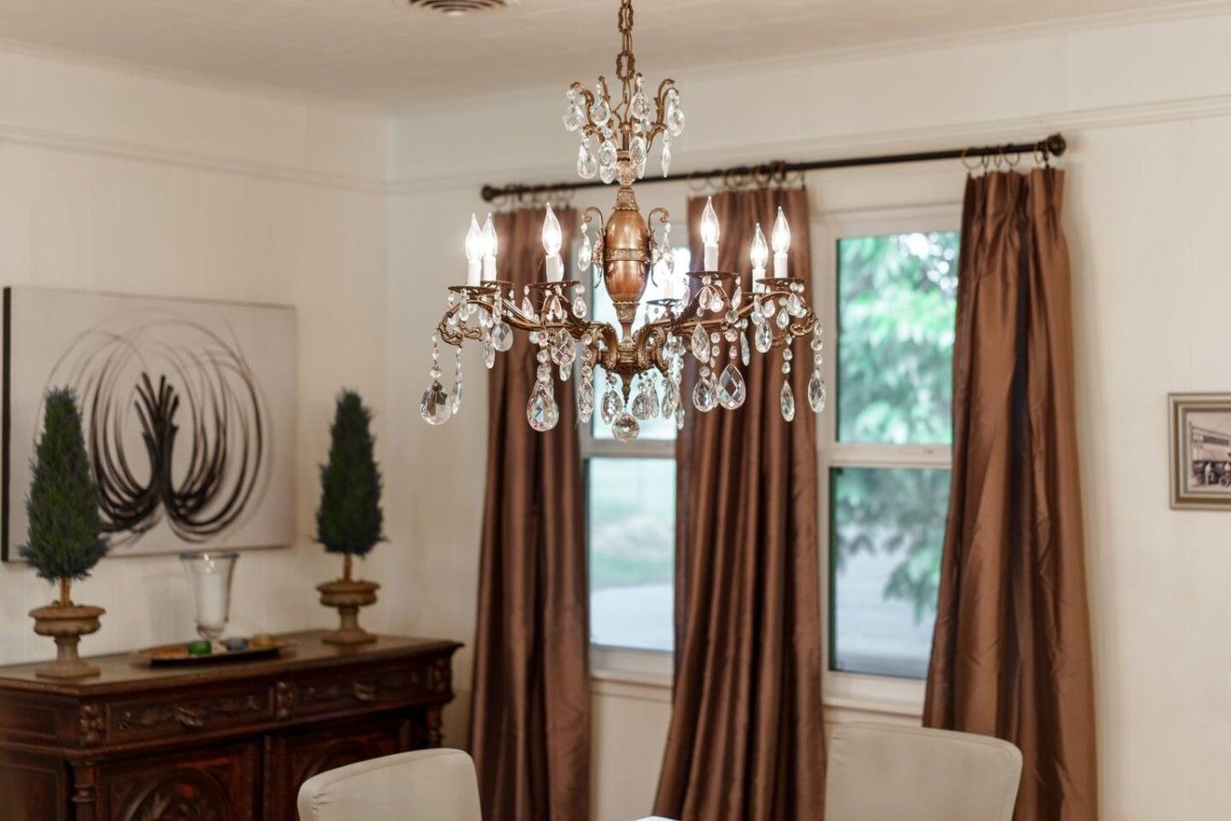 A dining area with a chandelier, brown curtains, a wooden cabinet, and a painting on the wall.