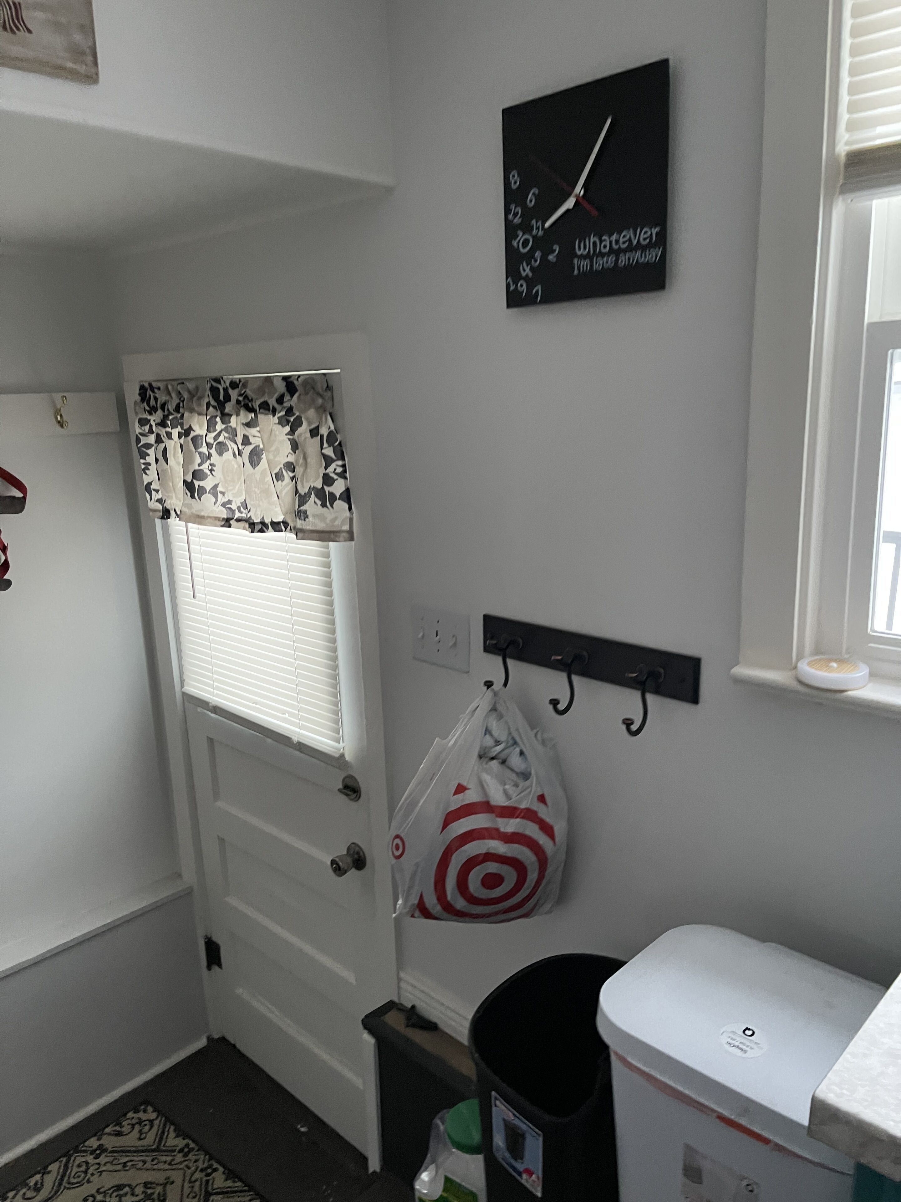 A small, clean room with a white door, a window with blinds, a clock on the wall, a Target bag hanging on the wall, and a trash can.