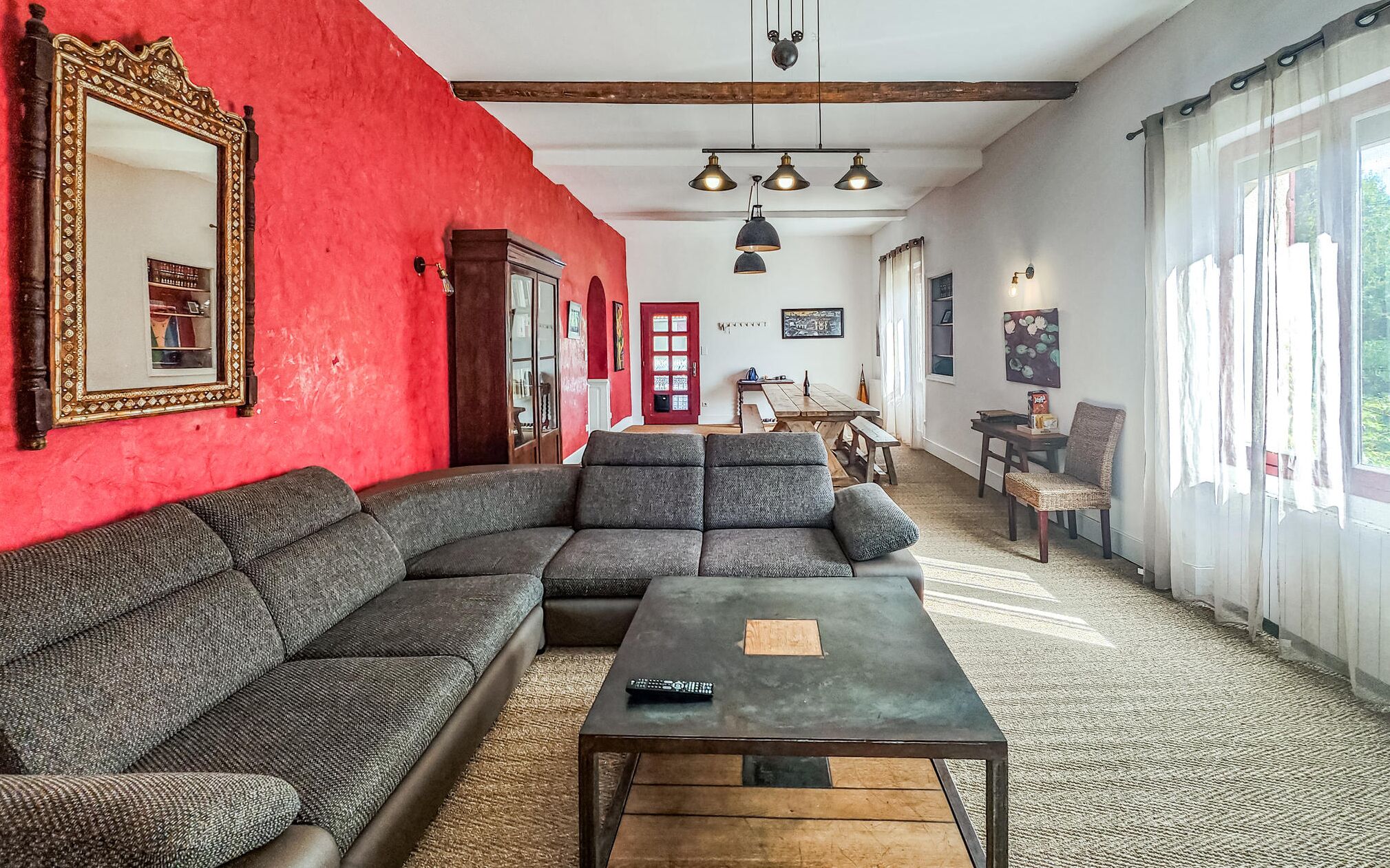 A living room with a red wall, a large mirror, a sectional sofa, a coffee table, and a dining area with a table and chairs.
