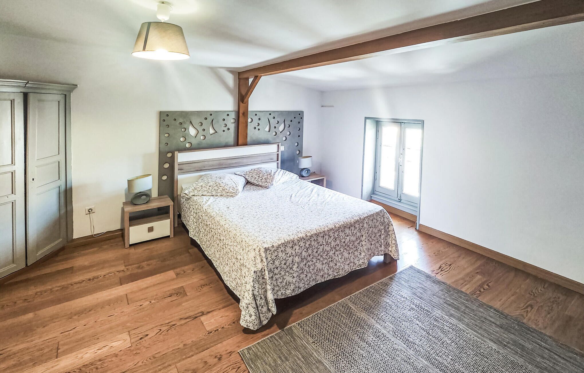 Une chambre avec un plancher en bois, un lit recouvert d’une couvre-lit à motifs floraux, une table de chevet avec une lampe, une étagère fixée au mur et une fenêtre avec des stores.