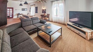 Un salon moderne comprenant un canapé d’angle, une table basse, un téléviseur posé sur un meuble en bois, ainsi qu’un coin repas avec une table et des chaises.