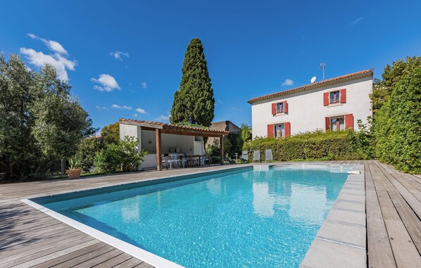 Ein Schwimmbad mit Holzboden, eine überdachte Terrasse mit Gartenmöbeln und ein zweistöckiges Gebäude mit roten Fensterläden.