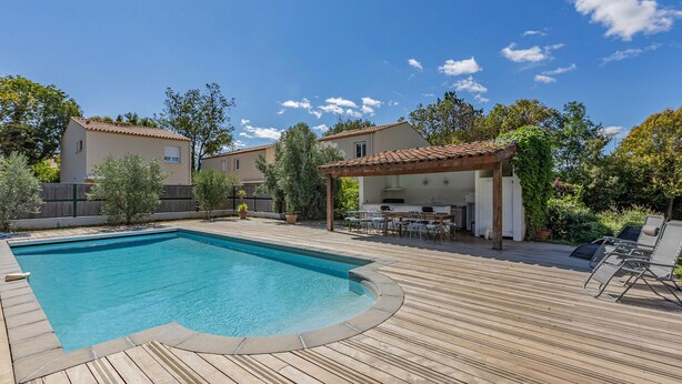 Une piscine avec une terrasse en bois, un espace repas extérieur couvert et deux maisons en arrière-plan.