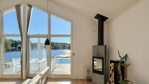 Amplia habitación con grandes ventanales con vistas a un lago, chimenea y vista a un muelle con mobiliario exterior.