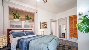 A bedroom with a bed, bedside tables, a nightstand, a mirror, a wardrobe, and a potted plant.