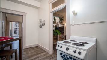 Een keuken met een witte kookplaat, een houten tafel en stoelen. Er is een deur naar een gang met een plant en een plank.