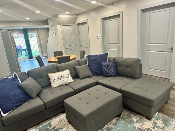 Ein graues Ecksofa mit einem 'BE OUR GUEST'-Kissen, einem kleinen runden Tisch und Blick ins Freie durch eine Schiebetür.