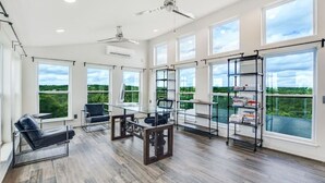 A spacious room with large windows, a ceiling fan, a black leather sofa, a small table, and a bookshelf. - 40-Acre Private Ranch  Pool  2 Homes Near Austin.  (Cedar Creek)