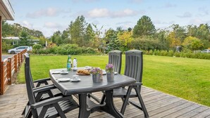 Un área de comedor al aire libre con mesa y sillas sobre una terraza de madera, rodeada por un césped y árboles.