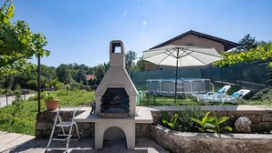 Une terrasse extérieure avec un barbecue en béton, un parasol blanc et un espace piscine équipé de chaises longues.