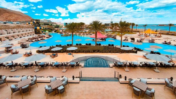 Un complex turístic amb una gran piscina, nombroses gandules, ombrel·les i vistes al mar.