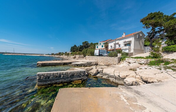 Une propriété située sur la côte, avec un chemin de pierre menant à une piscine aux eaux cristallines, un bâtiment blanc doté d’un toit rouge et une vue sur la mer.