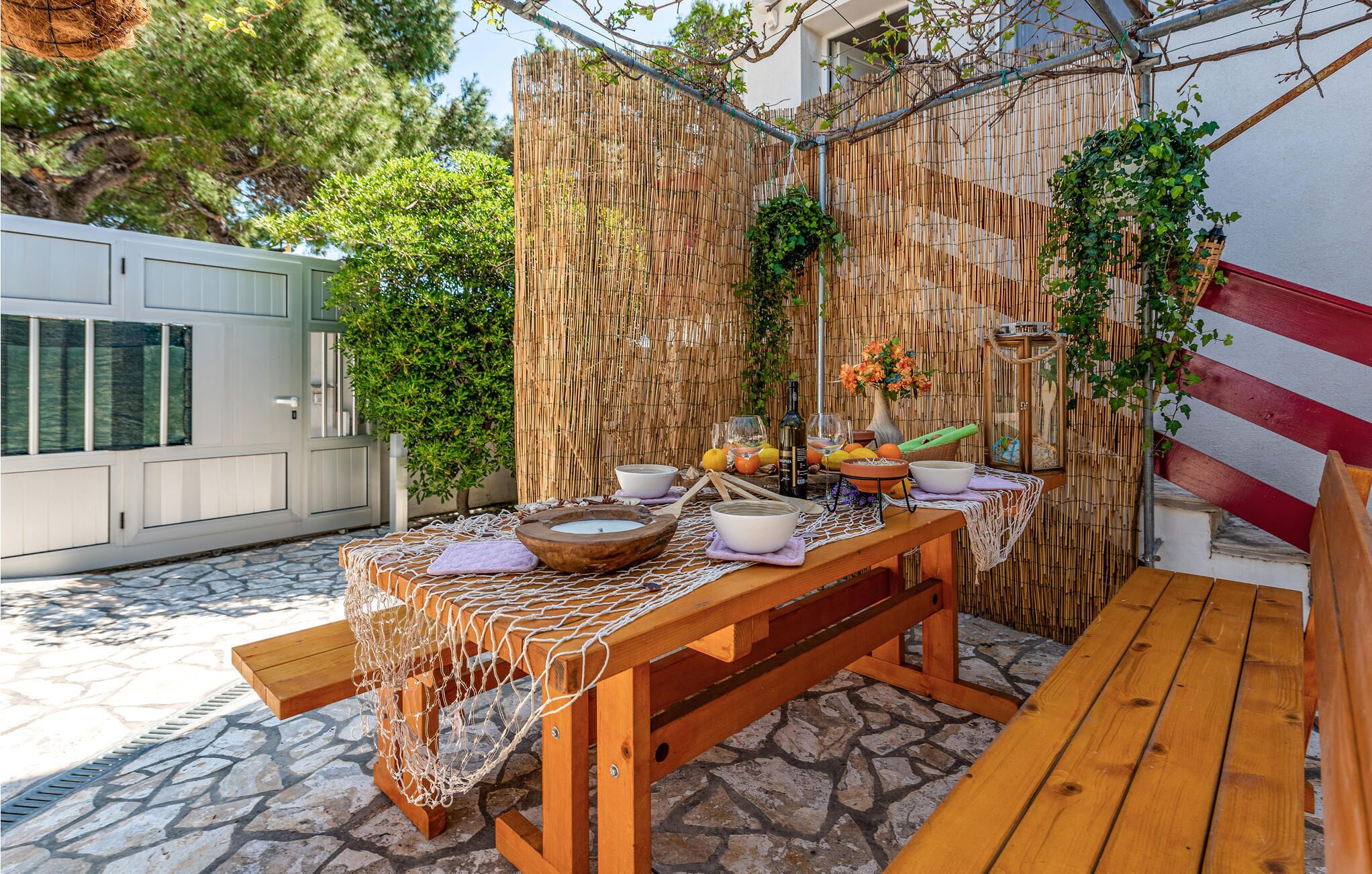 An outdoor dining area with a bamboo fence, a stone floor, and a wooden table set with bowls, glasses, and a bottle.