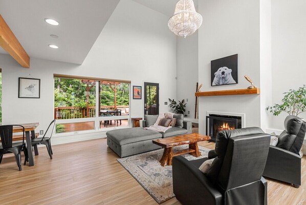 A modern living room with a fireplace, a dining area, and large windows overlooking a deck.