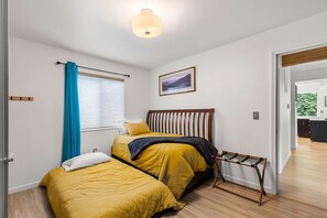 A bedroom with two beds, a wooden headboard, a window with blue curtains, and a view into the kitchen.