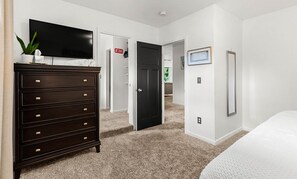 A bedroom with a dark wooden dresser, a flat-screen TV, a bed, and a door leading to another room.