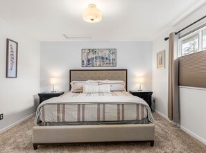 Un dormitorio con una cama grande, dos mesitas de noche, una lámpara en el techo, una ventana con persianas y una obra de arte en la pared.