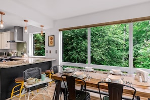 A modern kitchen with a large island, dining table set for four, and a view of lush greenery through large windows.