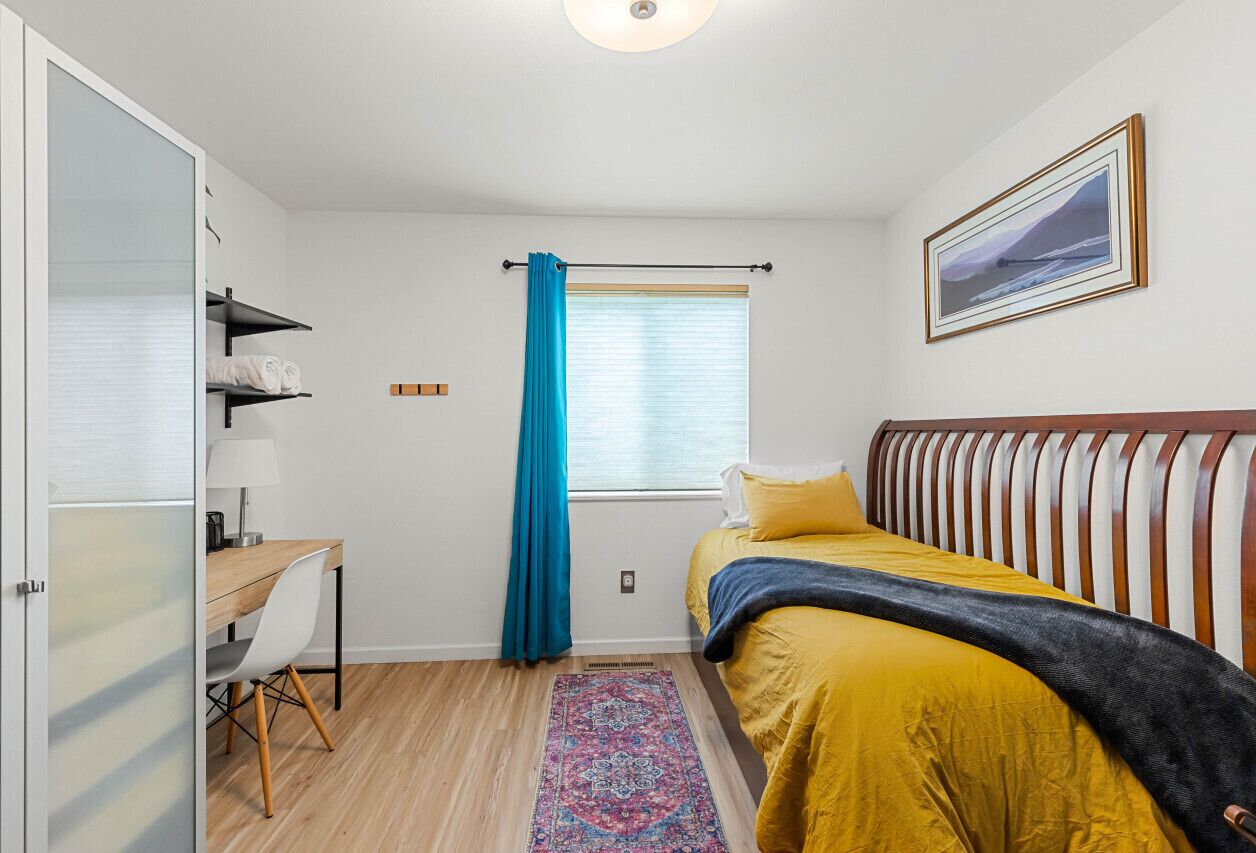 Ein Schlafzimmer mit einem Bett, einem Schreibtisch, einem Stuhl, einem Fenster mit Vorhängen und einem Bild an der Wand.
