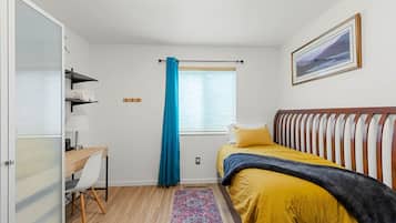 Un dormitorio con cama, escritorio, silla, ventana con cortinas y un cuadro en la pared.