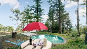 Une table en bois avec une bouteille et des verres, un parasol rouge et une piscine dans un espace boisé.