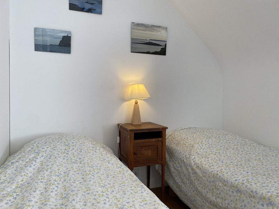 Una habitació petita i acollidora amb dos llits individuals, una tauleta de nit amb una làmpada i tres quadres a la paret.