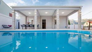 Un espace moderne aménagé au bord de la piscine, avec une piscine à l’eau d’un bleu limpide, des piliers en pierre et un espace détente couvert comprenant des chaises et une table.
