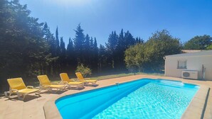 Une piscine rectangulaire à l’eau d’un bleu limpide, entourée d’une terrasse carrelée. Six chaises longues jaunes sont alignées le long du bord de la piscine. La zone de la piscine est adjacente à un bâtiment doté d’une fenêtre et d’un appareil de climatisation.