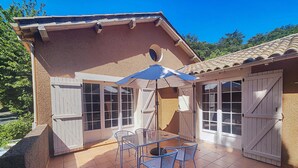 Une terrasse avec une table et des chaises, un parasol bleu et une vue sur un bâtiment doté de grandes fenêtres et d’un toit recouvert de tuiles.