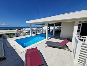 Uma área de piscina no terraço com vista clara para o oceano, um espaço de estar coberto e uma estrutura de edifício branca.