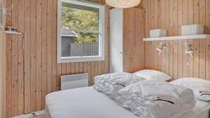 Un dormitorio con paredes de madera, una cama con ropa blanca, una ventana, una lámpara de techo y un radiador blanco.